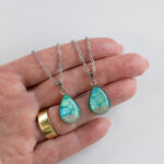 Ogrlica Soča v obliki kapljice, zeleno-modra z drobnim peskom iz reke Soče, ročno izdelana iz nerjavečega jekla. Prikaz v roki.