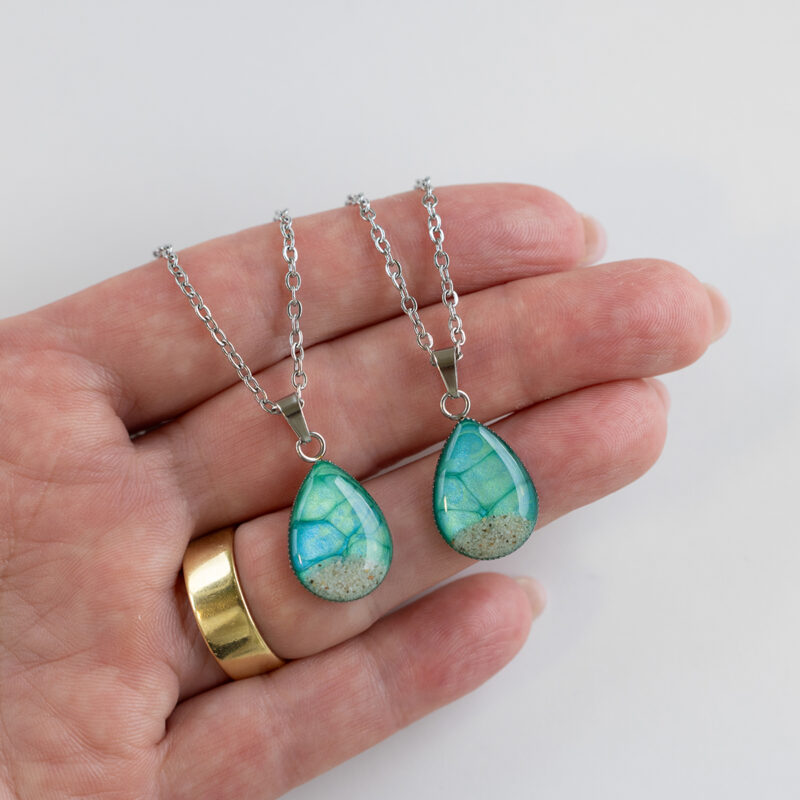 Ogrlica Soča v obliki kapljice, zeleno-modra z drobnim peskom iz reke Soče, ročno izdelana iz nerjavečega jekla. Prikaz v roki.