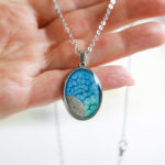 Ovalna ogrlica iz kolekcije Soča z modro-zelenim motivom in peskom iz reke Soče, v roki