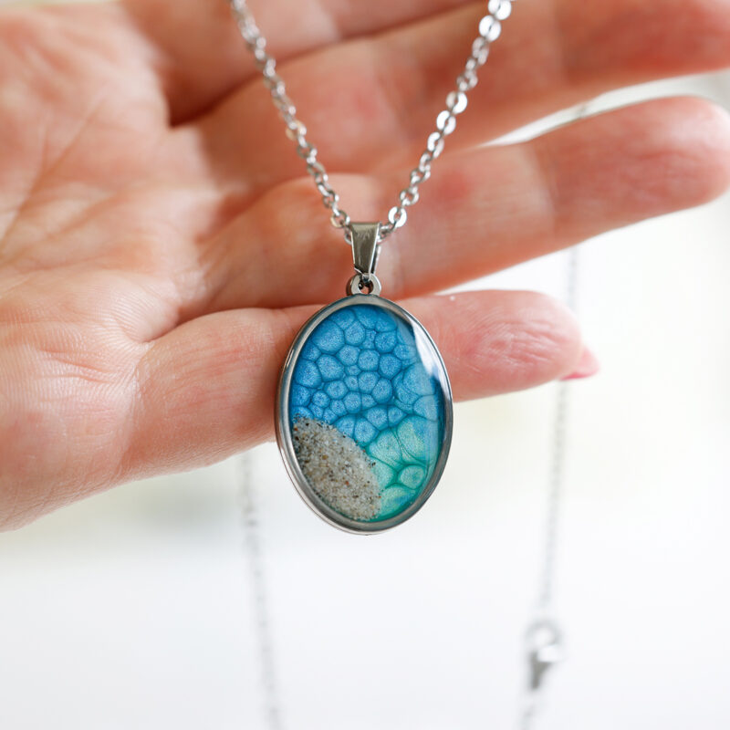 Ovalna ogrlica iz kolekcije Soča z modro-zelenim motivom in peskom iz reke Soče, v roki
