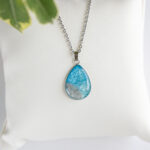 Ročno izdelana ogrlica s kapljičastim obeskom Soča v svetlo modri barvi s peskom iz reke, na beli podlagi.