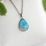 Ročno izdelana ogrlica s kapljičastim obeskom Soča v svetlo modri barvi s peskom iz reke, na beli podlagi.