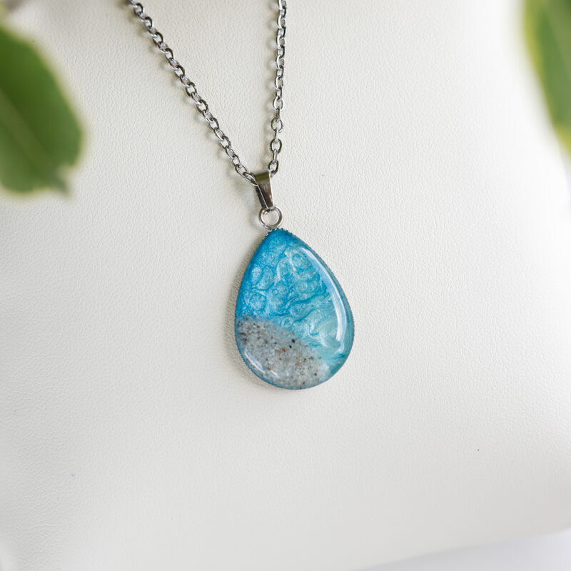 Ročno izdelana ogrlica s kapljičastim obeskom Soča v svetlo modri barvi s peskom iz reke, na beli podlagi.