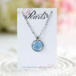 Ogrlica z več modrimi spominčicami v prozorni smoli in srebrnem okroglem obesku z verižico iz nerjavečega jekla – na kartončku Mountain Pearls