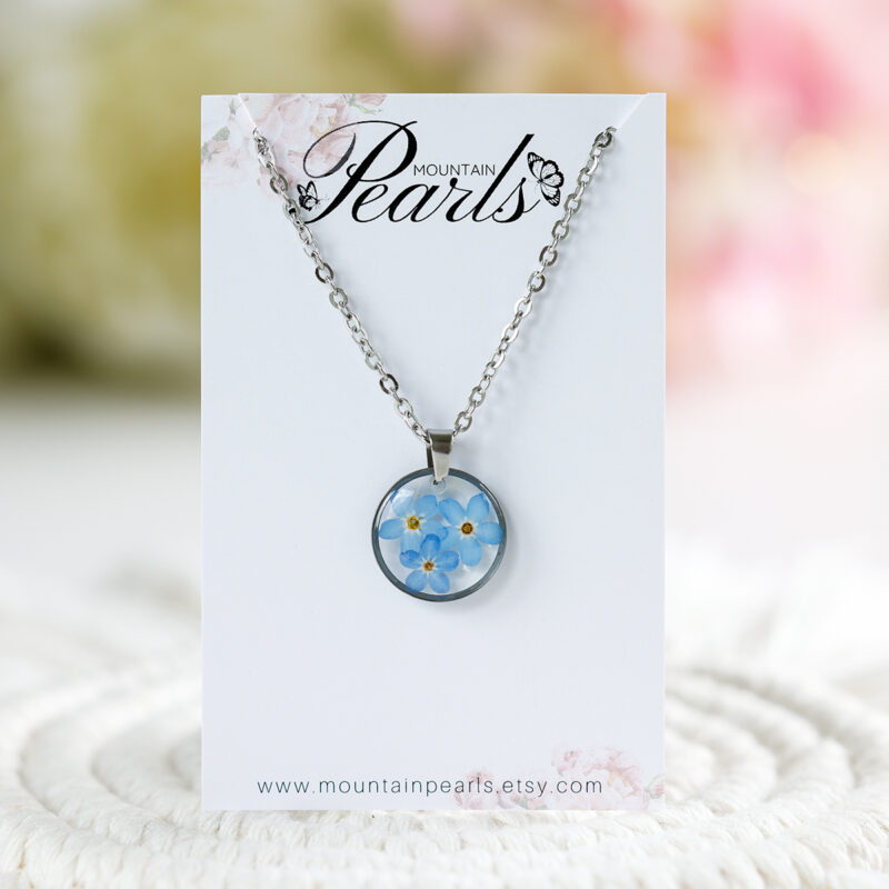 Ogrlica z več modrimi spominčicami v prozorni smoli in srebrnem okroglem obesku z verižico iz nerjavečega jekla – na kartončku Mountain Pearls