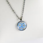 Ogrlica z več modrimi spominčicami v prozorni smoli in srebrnem okroglem obesku z verižico iz nerjavečega jekla.