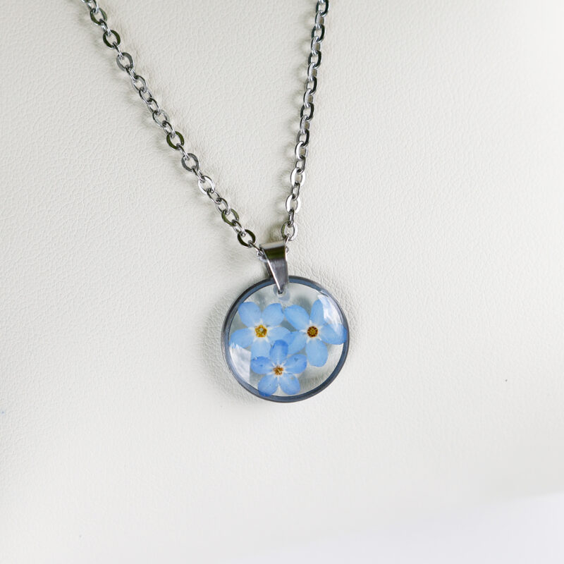 Ogrlica z več modrimi spominčicami v prozorni smoli in srebrnem okroglem obesku z verižico iz nerjavečega jekla.