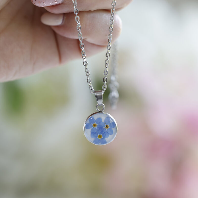 Ogrlica z okroglim obeskom, v katerem so ujete prave modre spominčice, premera 1,5 cm, prelita s smolo, prikazana v roki.