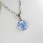 Ogrlica z okroglim obeskom, v katerem so ujete prave modre spominčice, premera 1,5 cm, prelita s smolo, prikazana na beli podlagi.