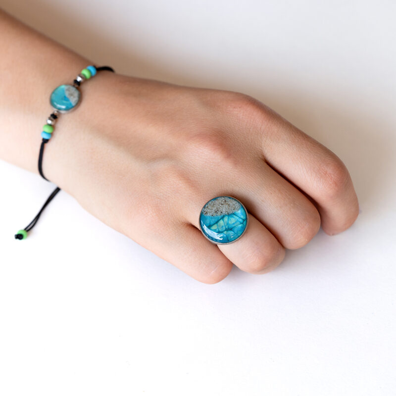 Ročno izdelan prstan Soča v turkizno-zelenih tonih z motivom peska iz reke Soče, premera 2 cm, na roki.