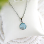 Svetlo modra ogrlica z okroglim obeskom iz kolekcije Soča, z dodatkom pravega peska in smolnim premazom, prikazana na beli podlagi.