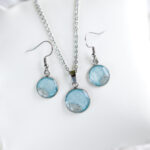 Svetlo moder komplet z ogrlico in uhančki iz kolekcije Soča, ročno pobarvan in z dodatkom pravega peska iz reke Soče, prikazan na belem ozadju.