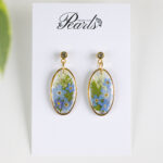 Ročno izdelani ovalni uhani s pravimi posušenimi modrimi in belimi cvetovi, ujeti v smolo, na jekleni osnovi.