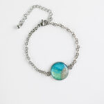 Ročno izdelana okrogla zapestnica z modro zelenim medaljonom in peskom iz reke Soče, premera 18 mm – lahkoten kos za ljubiteljico narave.